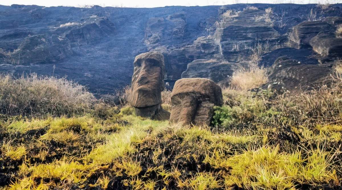 Two stone moai are visible in the foreground. In the background, there's a large black patch after a fire on Easter Island.