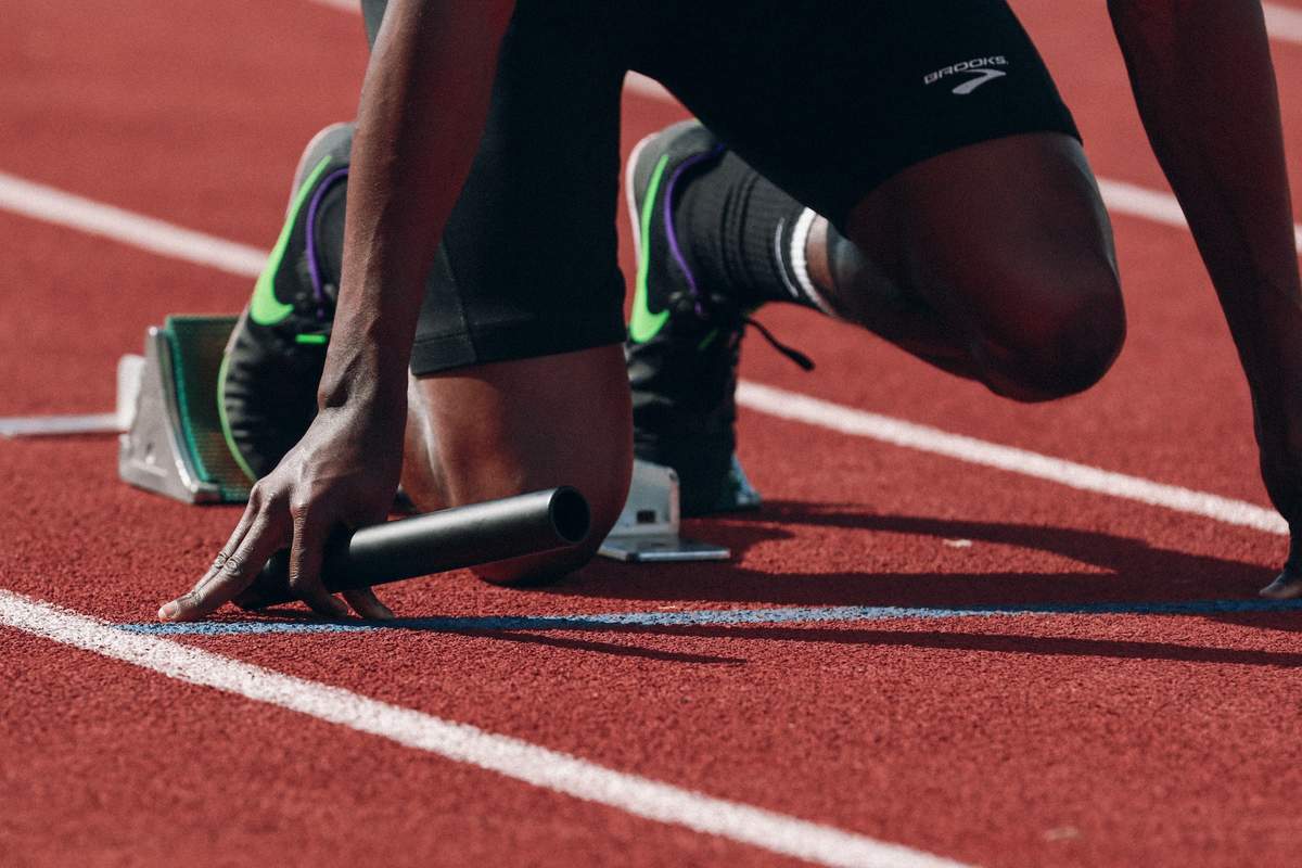 A runner with a baton prepares to begin a race on a track. Only the hands and legs of the runner are visible.