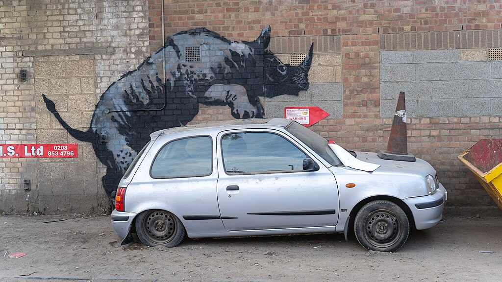 Banksy artwork - silhouette of a rhino who appears to be climbing on top of a crumpled car.