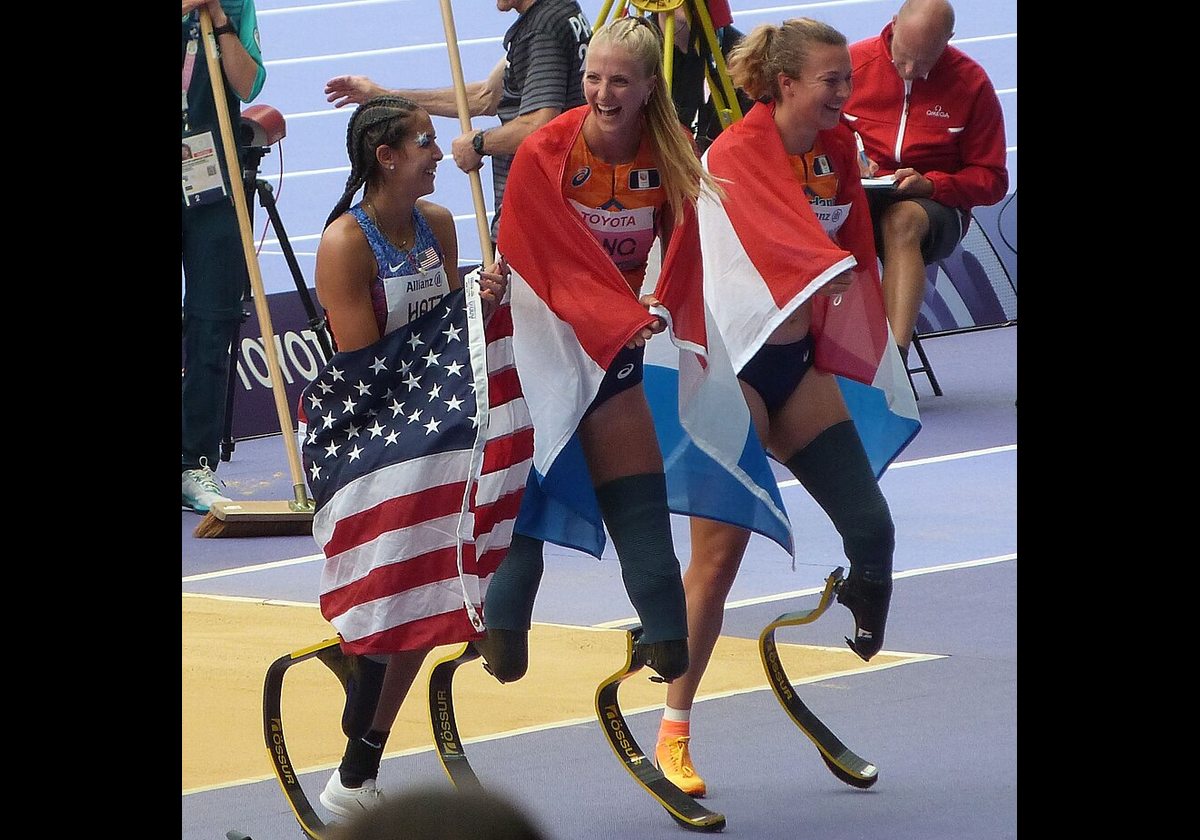 The three medal winners of the Women's long jump T64 stand together on their blade legs, each wrapped in the flag of their country during the 2024 Paris Summer Paralympic Games.